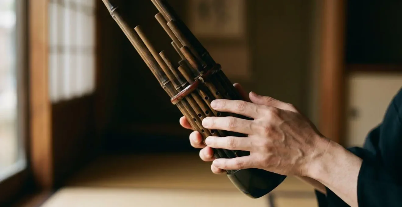 Human hands delicately covering finger holes on multiple Shō bamboo pipes to form an aitake chord, captured in soft natural light
